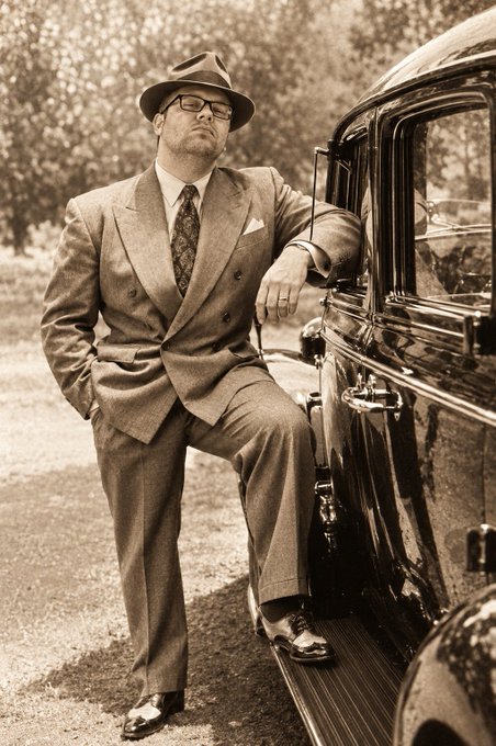A sepia-toned vintage photograph of a bearded man in a double-breasted suit and fedora, posing next to a classic automobile.