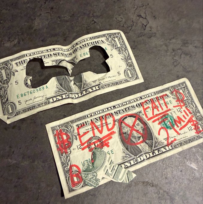 A pair of torn and defaced US one-dollar bills lying on a dark stone surface; the top bill has a face-sized hole, while the bottom bill is marked with red graffiti reading "LIES" and "AMERICA FAILS."