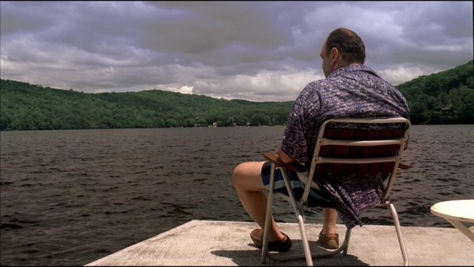 A man wearing a purple patterned shirt sitting on a folding chair on a wooden dock, looking out over a lake surrounded by green hills under a cloudy sky.