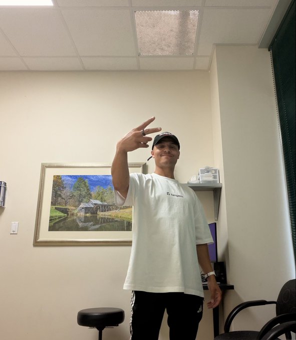 A person wearing a cap and white t-shirt posing with a peace sign in what appears to be a medical or office room with a landscape painting on the wall.
