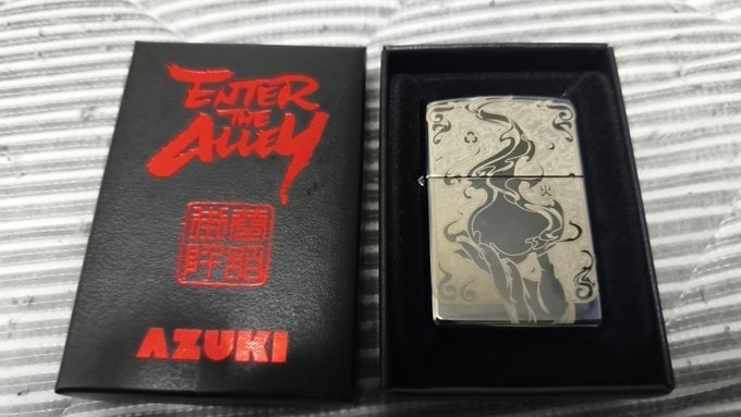 The image features a silver lighter with an Azuki design, placed inside its original black box. The box has a red label that reads "Enter the Alley" along with some Japanese characters. The lighter itself is silver and has a unique design featuring a woman's face on it.