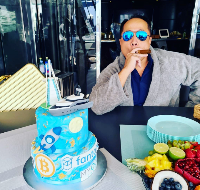 A man wearing blue sunglasses and a gray cardigan smokes a cigar behind an elaborate blue birthday cake decorated with logos for Bitcoin, Fantom, and "MYC," alongside a yacht topper, while fresh fruit plates sit on the table. The image reflects the post's context of a wealthy early cryptocurrency investor, contrasting symbols of crypto fortune and luxury with the tragic fate mentioned in the caption.