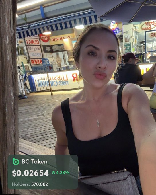 A woman taking a selfie at an outdoor location with shop signs in the background, overlaid with a green ticker showing BC Token price at $0.02654, up 4.28%, with 570,082 holders.