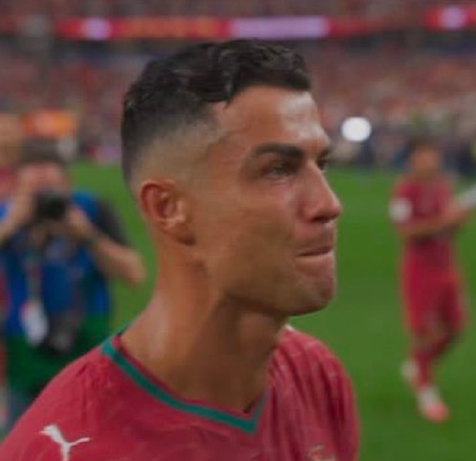 A close-up of Cristiano Ronaldo in his red Portugal kit looking to the side on the pitch, with blurred stadium lights and a teammate visible in the background.