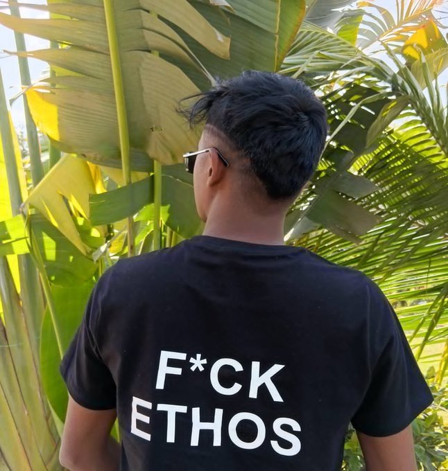 A photo of a young man from behind, wearing sunglasses and a black t-shirt with the text 'F*CK ETHOS', standing in front of tropical foliage.