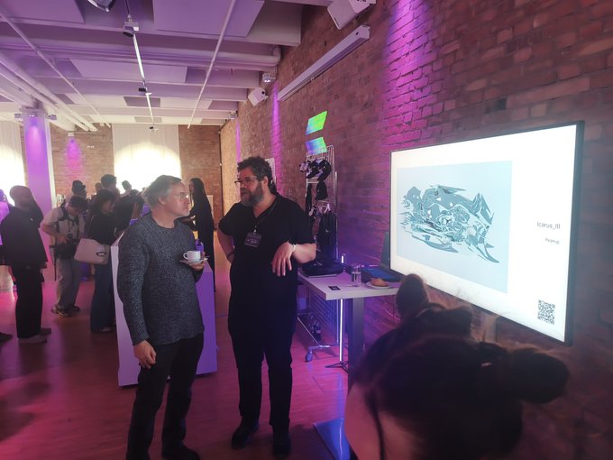 The image shows a group of people gathered around a television screen displaying an art exhibit, with two men standing close together and engaged in conversation. The room has a brick wall behind them, and there are other people present as well. A few individuals can be seen holding drinks or coffee cups, suggesting that they might be enjoying refreshments while appreciating the artwork on display.