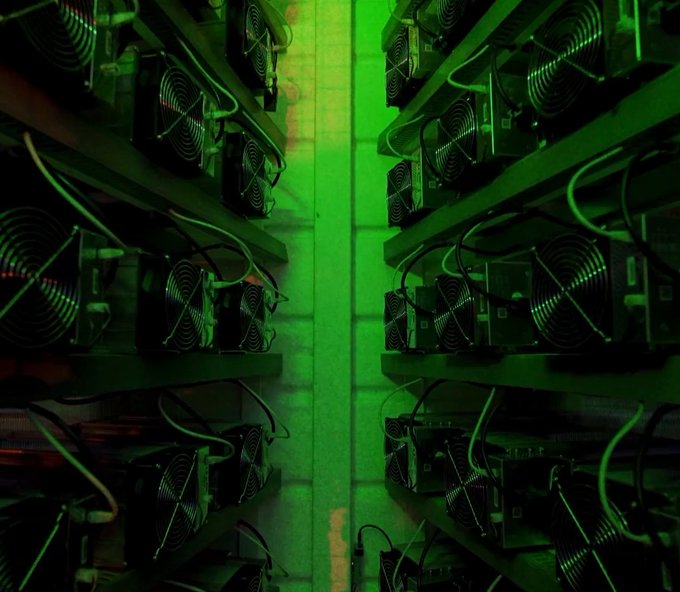 Rows of electronic mining rigs with cooling fans line both sides of a narrow aisle, illuminated by a green light.