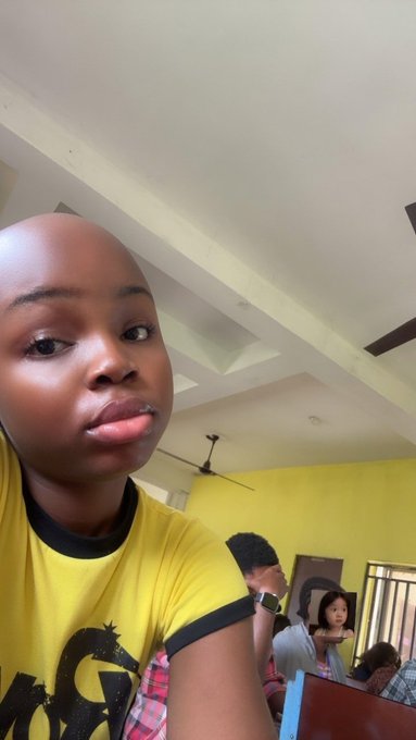 A young woman with a shaved head takes a selfie while sticking out her tongue, wearing a yellow shirt with black trim, in what appears to be a home or classroom setting with a ceiling fan and yellow walls visible behind her. The casual selfie fits the morning greeting vibe of the post wishing followers "gm" (good morning) and encouraging them to "have a good day."