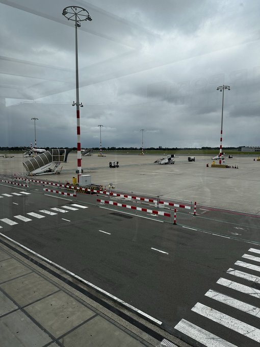 A view from inside an airport terminal shows the tarmac under overcast gray skies, with red and white light poles, a covered walkway, and ground equipment visible on the runway. The cloudy weather outside the window perfectly matches the post's sentiment about leaving gloomy skies behind for a sunnier destination.