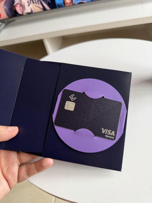 A hand holding an opened dark blue presentation folder containing a black Visa Signature metal card mounted on a purple circular card holder.