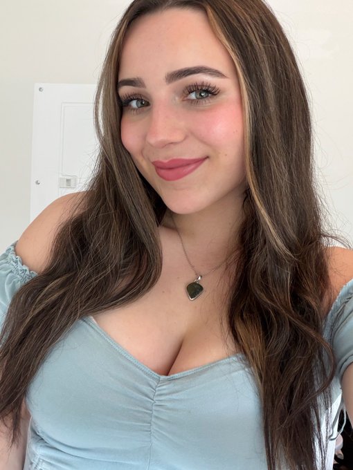 A woman with long brown hair wearing a light blue off-the-shoulder top and a heart pendant necklace poses for a selfie, complementing the caption's playful message about her future husband struggling financially while she's waiting for him.