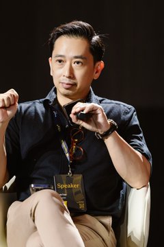 A man with dark hair wearing a dark button-up shirt, sitting on a chair and speaking with hand gestures, wearing a 'Speaker' lanyard.