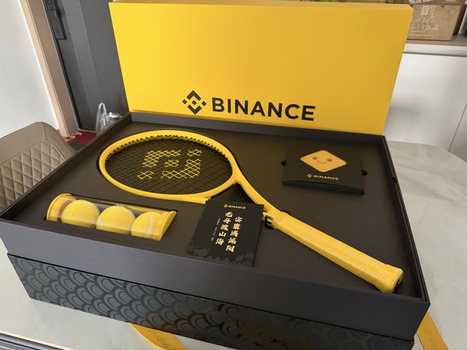 The image features a tennis racket, tennis balls, and a box containing these items. The tennis racket has yellow strings and a black handle. There are three tennis balls inside the box along with a small card that says "币友们端午安康". The post in the image is about a new product from Binance, which includes a tennis racket and tennis balls as part of its offering.

The main focus of the image is on the tennis equipment, while the background shows a dining table with chairs around it. This suggests that the scene might be taking place at a restaurant or a similar setting where people can enjoy their meals together.