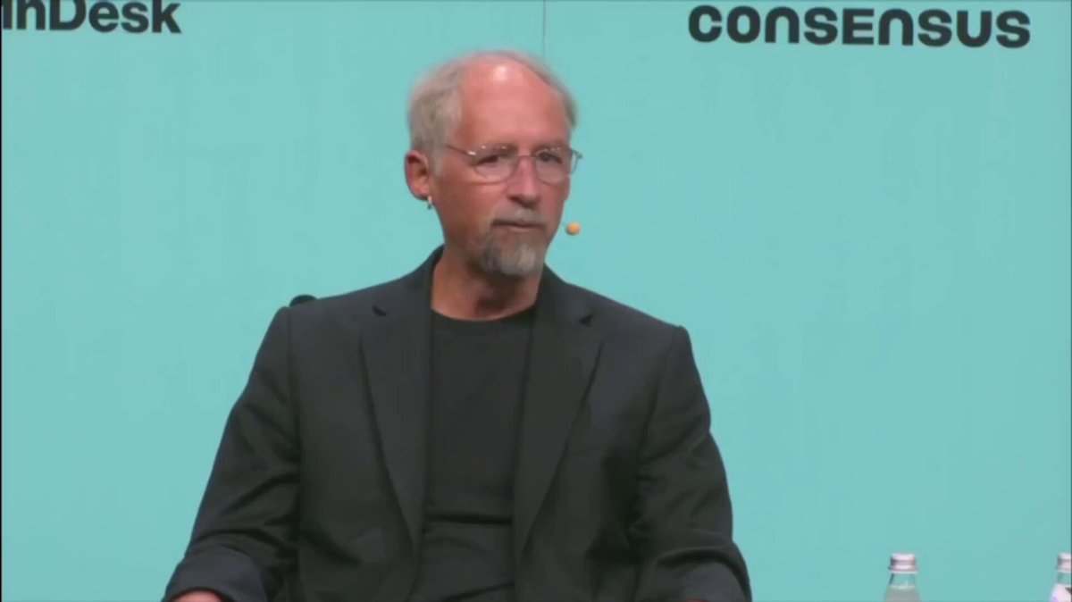 The image shows a man wearing glasses, sitting at a desk with his hands crossed. He appears to be engaged in a conversation or presentation, possibly discussing business strategies. The background features a blue wall that has the words "consensus" written on it. There are also two bottles placed near the edge of the frame and a water bottle closer to the center of the image.