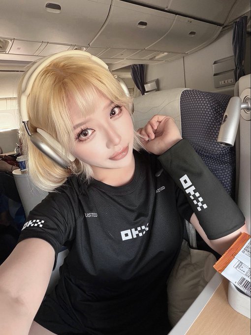 Selfie of a blonde woman wearing headphones and a black athletic shirt with 'OKX' branding, sitting in what appears to be an airplane seat.