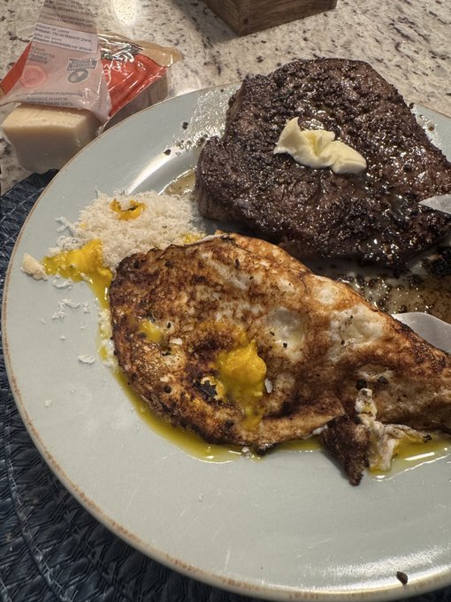 A breakfast spread of a dark-crusted steak with butter, alongside crispy eggs over rice on a gray plate, posted with the caption "GM   what's cooking today???". The image showcases a hearty meal being shared in response to the greeting about what's on the menu.