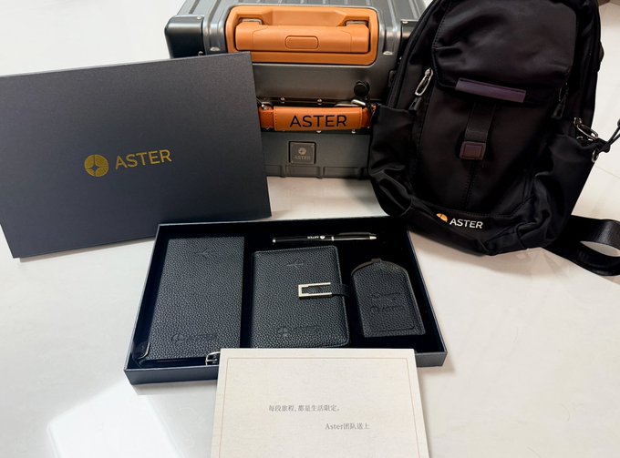An Aster DEX branded gift set is displayed, featuring a black box with the gold "ASTER" logo, a matching orange and gray carry-on suitcase, a black backpack, a passport holder, notebook, and other accessories presented in a tray with an "Aster" welcome card in the foreground. The image showcases the premium travel merchandise mentioned in the post, gifted to aster-2:native holders as promotional items for the Aster community.