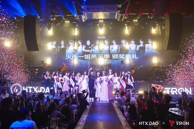The image shows a group of people standing together for an event at a concert or festival. The crowd consists of many individuals wearing white outfits, with some holding signs that say "HTX DAO" and "From the Huobi Sister Stage to the HTX DAO Community, I'm in I'm Xiao Du, HTX DAO @HTX_DAO Community Ambassador Many friends first got to know me from the "Huobi Sister" competition stage. The event is taking place at a venue with a large screen and speakers, creating an atmosphere of excitement and celebration.