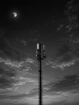 A black-and-white photograph of a tall cellular tower silhouetted against a cloudy night sky with a crescent moon visible in the upper left.