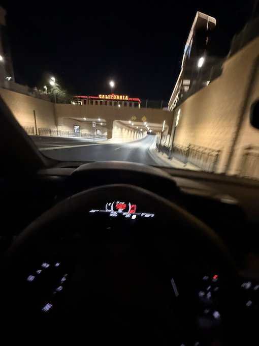 A nighttime view from the driver's seat of a car with its dashboard lights on, approaching the Bally's hotel in Las Vegas. The illuminated "BALLY'S" sign is visible in the distance, adding to the late-night Vegas atmosphere that matches the casual "Heh" reaction.