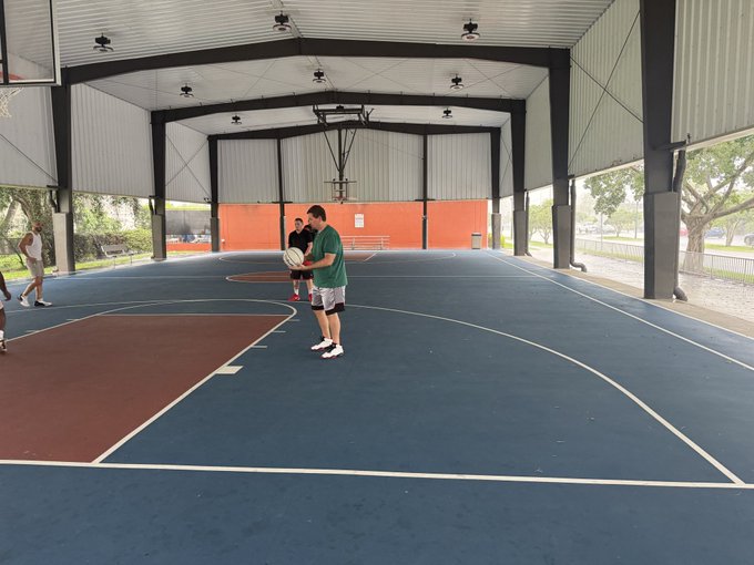 An indoor covered basketball court with a blue playing surface where a man in a green shirt holds a basketball, with another person visible in the background and a basketball hoop mounted overhead.