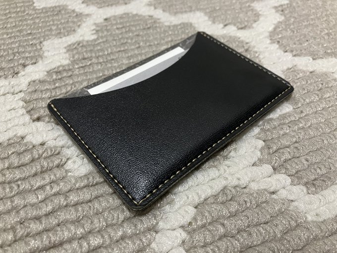 A black leather card holder with tan stitching rests on a grey and white patterned rug, with a white card partially visible in its slot.