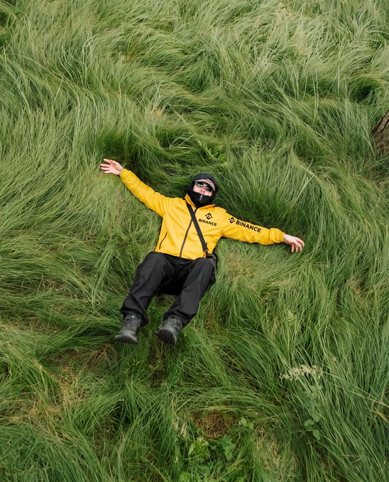 A person wearing a yellow jacket with "BINANCE" branding lies sprawled in lush green grass, arms outstretched as if embracing the wild field. The image playfully illustrates the caption "Grass the touch." by literally showing someone getting up close and personal with grass.
