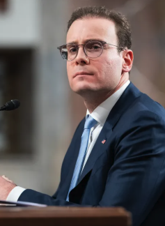CFTC Chair Mike Selig, wearing glasses and a dark suit with a light blue tie, is seen seated at a microphone, addressing crypto market structure and the CLARITY Act.