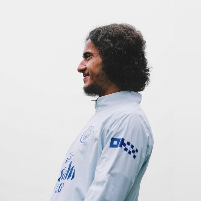 A man with curly hair and a beard smiles in profile against a pale background, wearing a light blue training jacket featuring the "OKX" sponsor logo and the Manchester City crest, aligning with the post's theme of unveiling new season kit and fresh faces in club apparel.