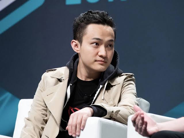 A young man with short dark hair, wearing a beige trench coat over a black hoodie, sits in a white chair during what appears to be an interview or panel discussion.