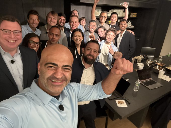 A group selfie of many smiling people in business attire gathered in a conference room, with a bald man in a blue shirt taking the photo in the foreground.