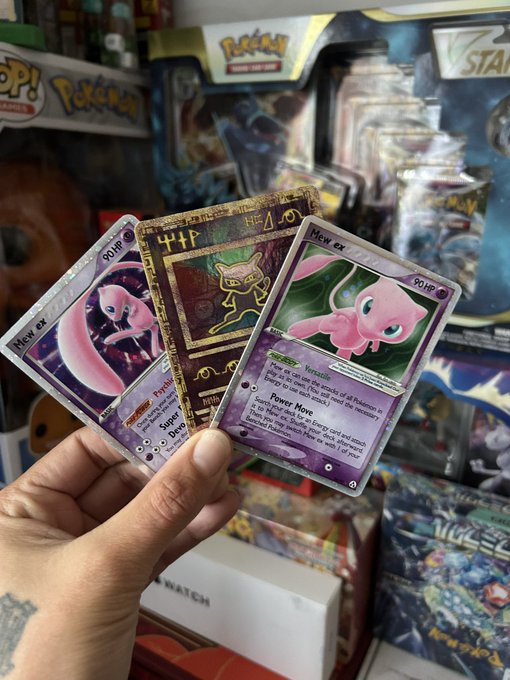 A hand holding two Mew ex Pokémon cards and a gold ancient Mew promo card, photographed in front of a display of Pokémon TCG merchandise including tins and blister packs.