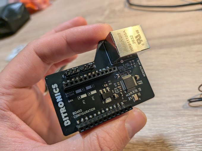A hand holds a small black electronic breakout board labeled 'BITRONICS BOARD CONFIGURATION' featuring a metal relay component marked 'HJR-4105-L-05V'.
