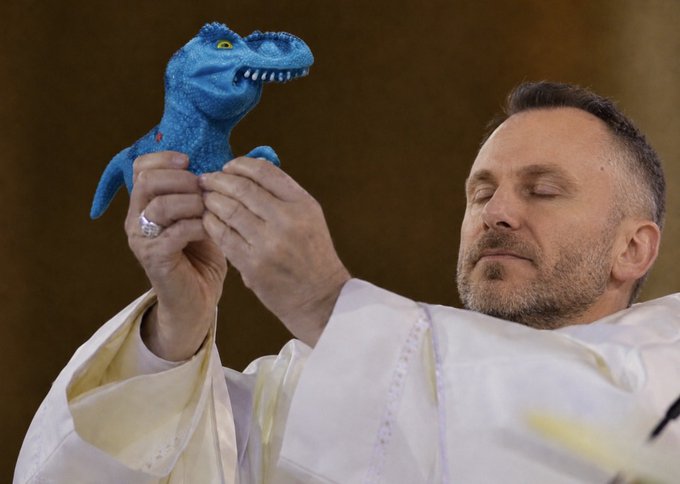 A man in white religious vestments holding up a small blue dinosaur puppet while his eyes are closed.
