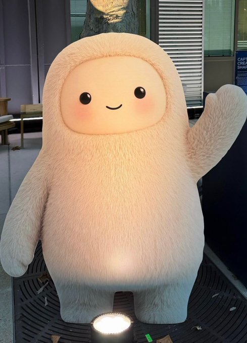 A large, fluffy white mascot statue with a smiling face, blushing cheeks, and a raised arm waves at the camera.
