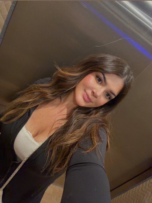 A selfie-style photo of a woman with long brown hair wearing a black jacket over a light top, standing near what appears to be a metallic surface.