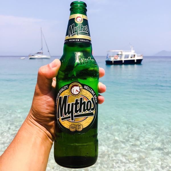 The image features a person holding up a green bottle of Mythos beer, which has a label with Greek text on it. The scene takes place at the beach, where boats can be seen in the background. The person's hand is positioned over the beer bottle, and they are likely enjoying their drink while taking in the beautiful view of the ocean.