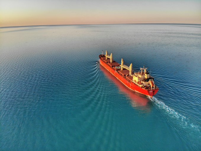 An aerial view of a red cargo ship sailing on calm blue waters at sunset or sunrise, leaving a wake behind it.
