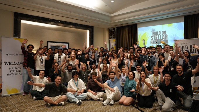 The image shows a large group of people gathered together for an event or celebration. They are posing for a photo with their arms raised, indicating a sense of accomplishment or excitement. The room they're in appears to be a conference hall or a similar venue, as there is a banner hanging on the wall behind them and a projector screen visible in the background.

The group consists of people from various age groups and genders, all dressed similarly with white shirts. They are standing close together, creating a sense of unity and camaraderie among them. The event seems to be focused on celebrating their achievements or milestones related to the hackathon they participated in.