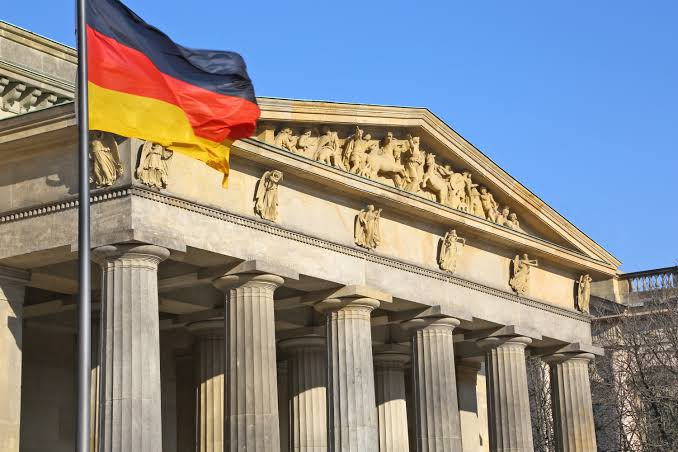 A photograph of the German national flag (black, red, and gold) waving in the foreground in front of a grand neoclassical stone building with tall columns and an ornate sculpted pediment under a clear blue sky.