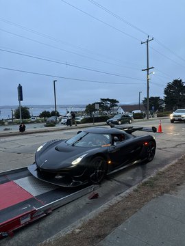 A sleek black Lamborghini sports car is being loaded onto a flatbed tow truck on a street near the ocean.