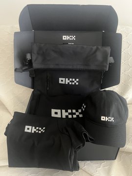 A collection of black OKX-branded merchandise including bags, a cap, and clothing arranged on a surface.