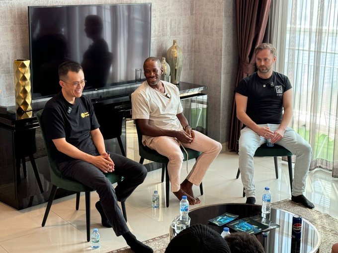 Three men sit on chairs in a modern living room setting for what appears to be a panel discussion or interview, with water bottles and pamphlets on a glass coffee table.