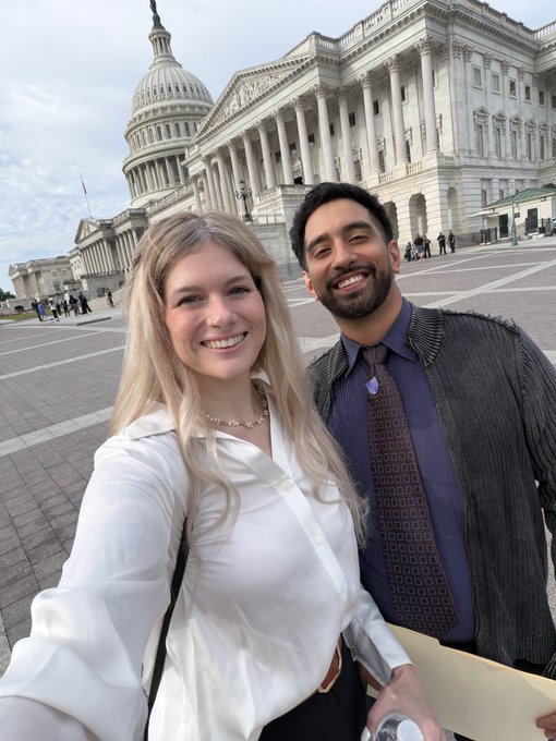 A selfie of a smiling blonde woman and bearded man standing in front of the United States Capitol building.