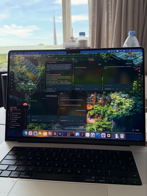 A MacBook displays a Slack workspace with multiple chat windows open, overlaid on a vibrant garden wallpaper. The setup sits on a desk near a window with a water bottle nearby, perfectly capturing the productivity-meets-aesthetic vibe of a freshly organized "Fable 5 vibe space."