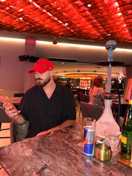 A man wearing a red cap sits at a bar table looking at his phone, with a hookah, Red Bull can, and champagne bottle in the foreground under a red-lit ceiling—fitting a casual "Gm friends" morning greeting.