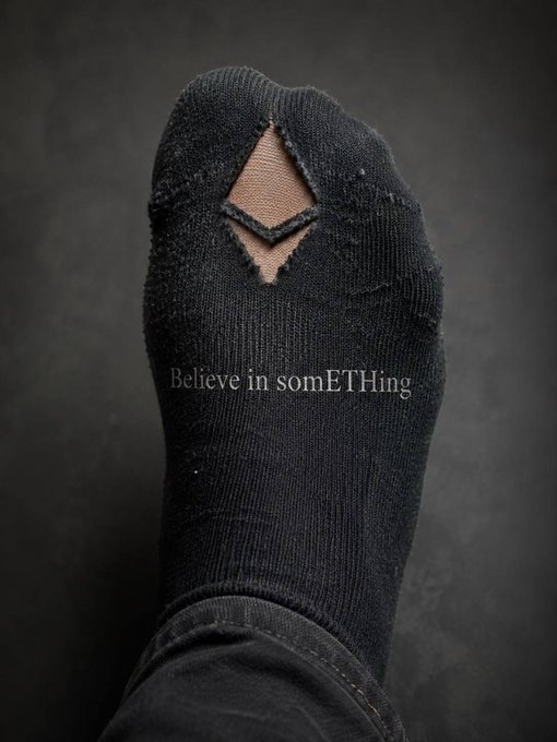 A foot wearing a worn black sock with a hole at the toe displays the text "Believe in somETHing," visually referencing Nike's iconic "Believe in something" slogan through the exposed skin forming the "E" in "something."