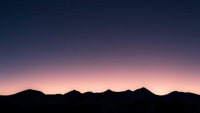 A serene landscape photograph showing the dark silhouettes of mountain ridges against a gradient twilight sky transitioning from pinkish-orange at the horizon to deep purple above.