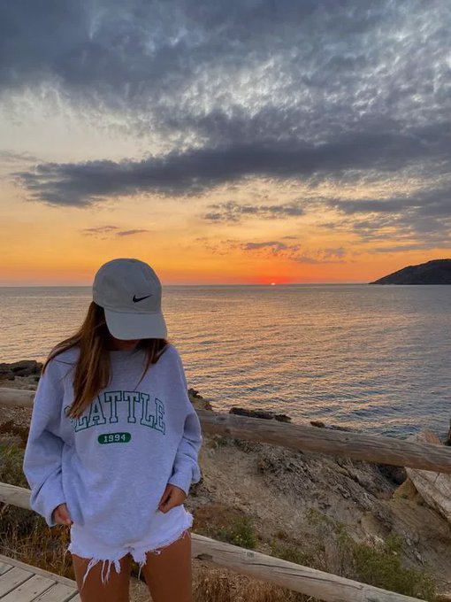 A woman in a Nike cap and grey "Seattle 1994" sweatshirt stands at a coastal viewpoint as the sun sets over the ocean, her face hidden as she looks down. The serene, early-stage sunset evokes catching a story just as it begins — quietly positioning ahead of the crowd before the rest of the sky catches on.