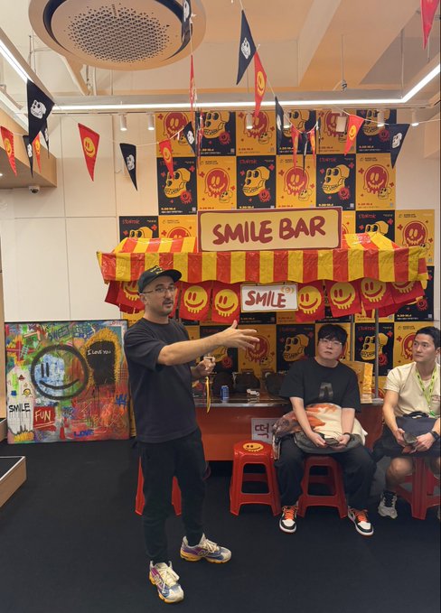 A man in a black shirt and cap gestures while speaking in front of a 'SMILE BAR' booth decorated with skull-themed artwork, red and yellow flags, and a striped awning, with two other people seated nearby.